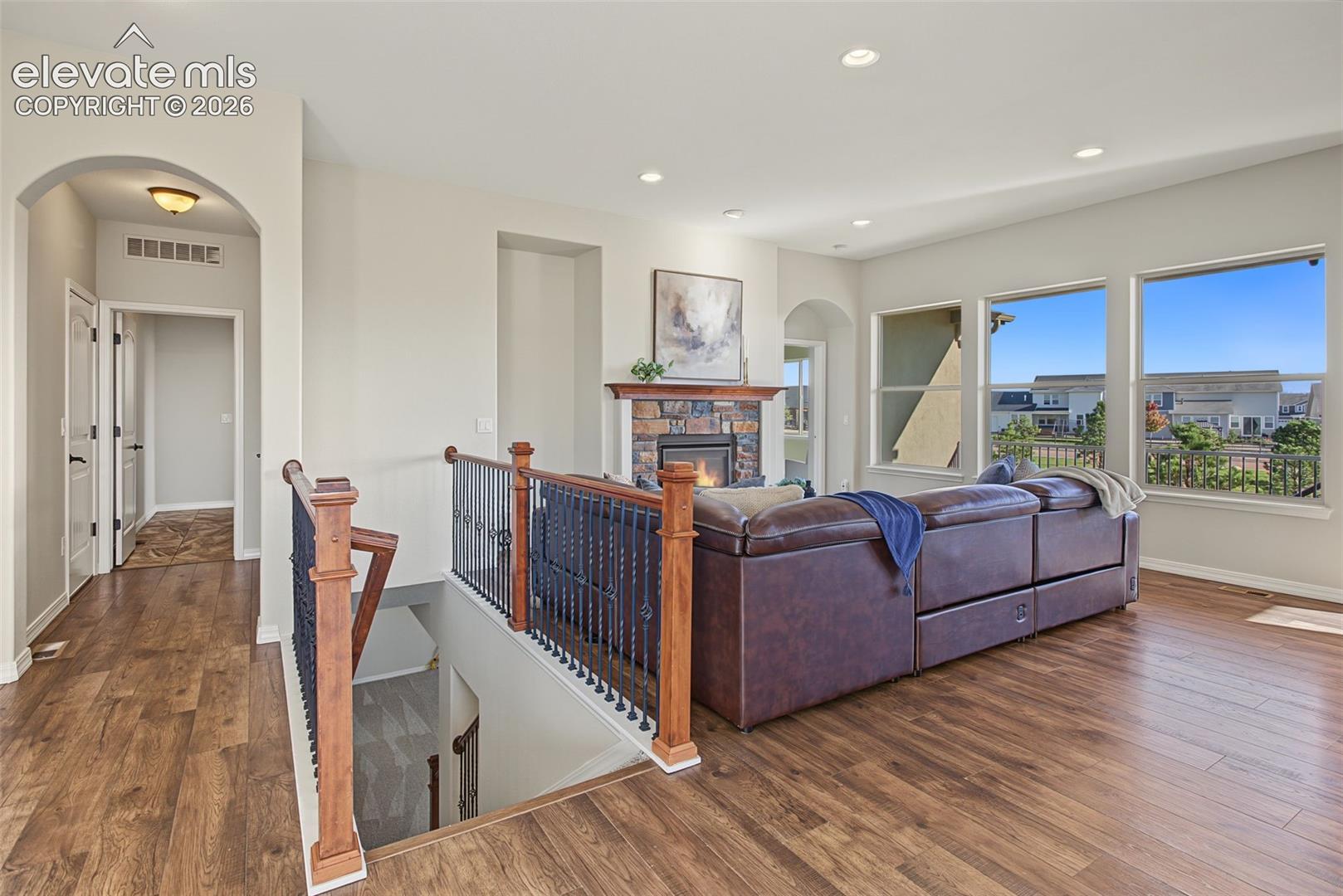 Wood-finish flooring, stone-surround fireplace, wrought-iron baluster railing, arched architectural detailing, and recessed lighting