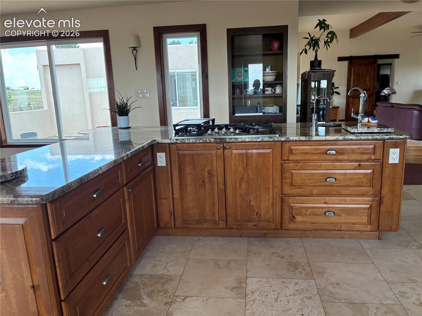 Kitchen island featuring a gas cooktop, integrated sink with gooseneck faucet, light granite countertops, and wood cabinetry with brushed nickel hardware