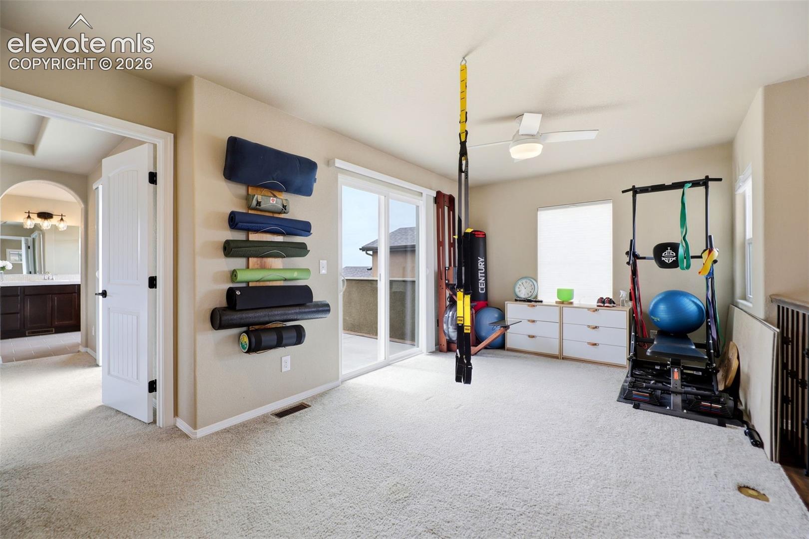 Carpeted loft being used as an exercise room, with a ceiling fan, sliding glass door leading out to the upstairs balcony