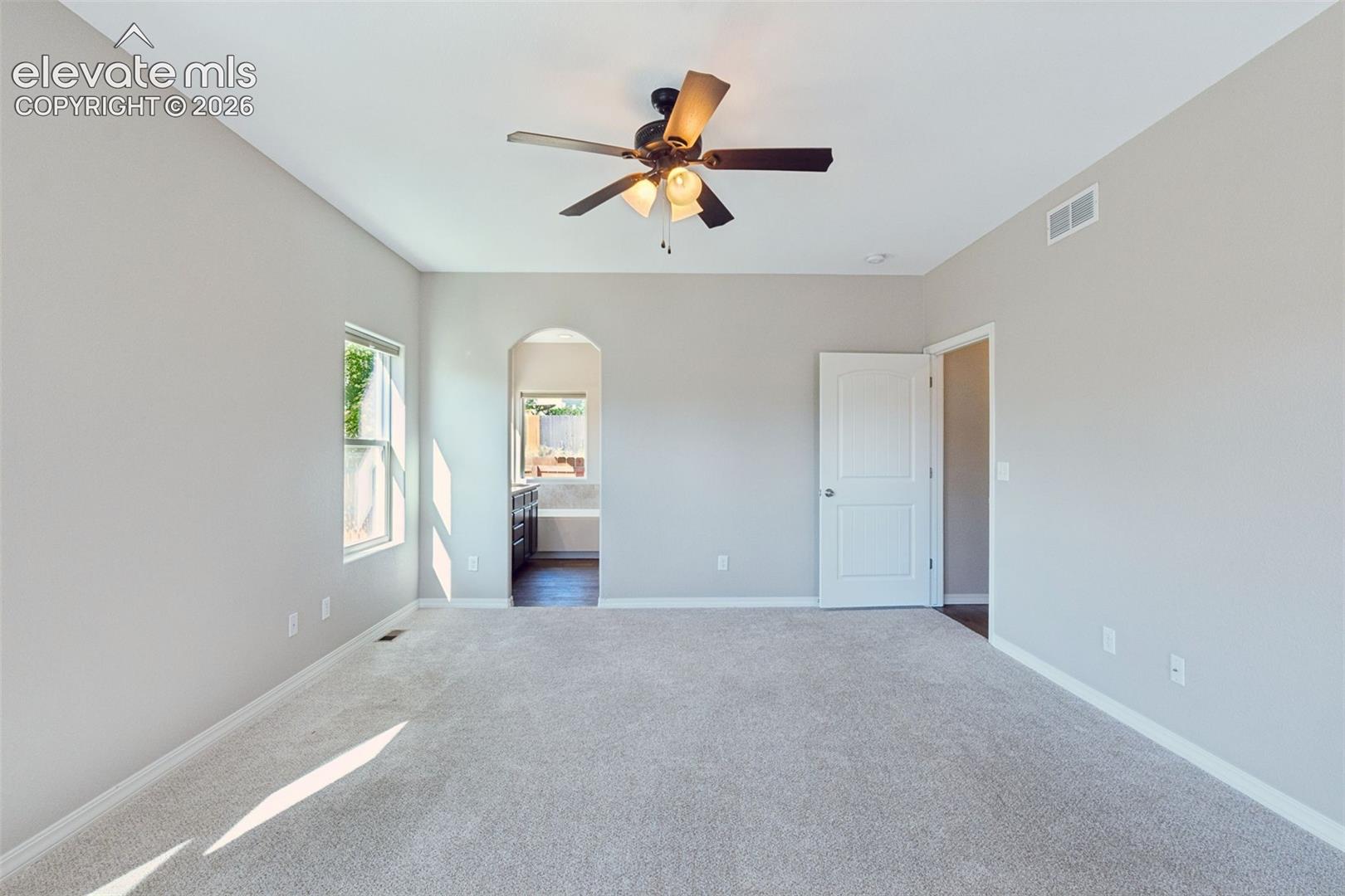 Spacious room featuring light gray walls, plush carpeting, a ceiling fan with integrated lighting, a window, and an arched doorway leading to an additional space