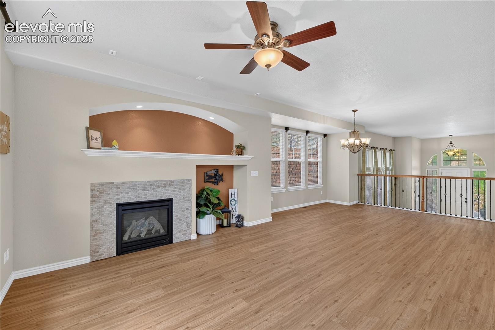 Fireplace with stone-tile surround, arched recessed shelving, wood-finish flooring, ceiling fan with integrated lighting, and decorative metal stair railing