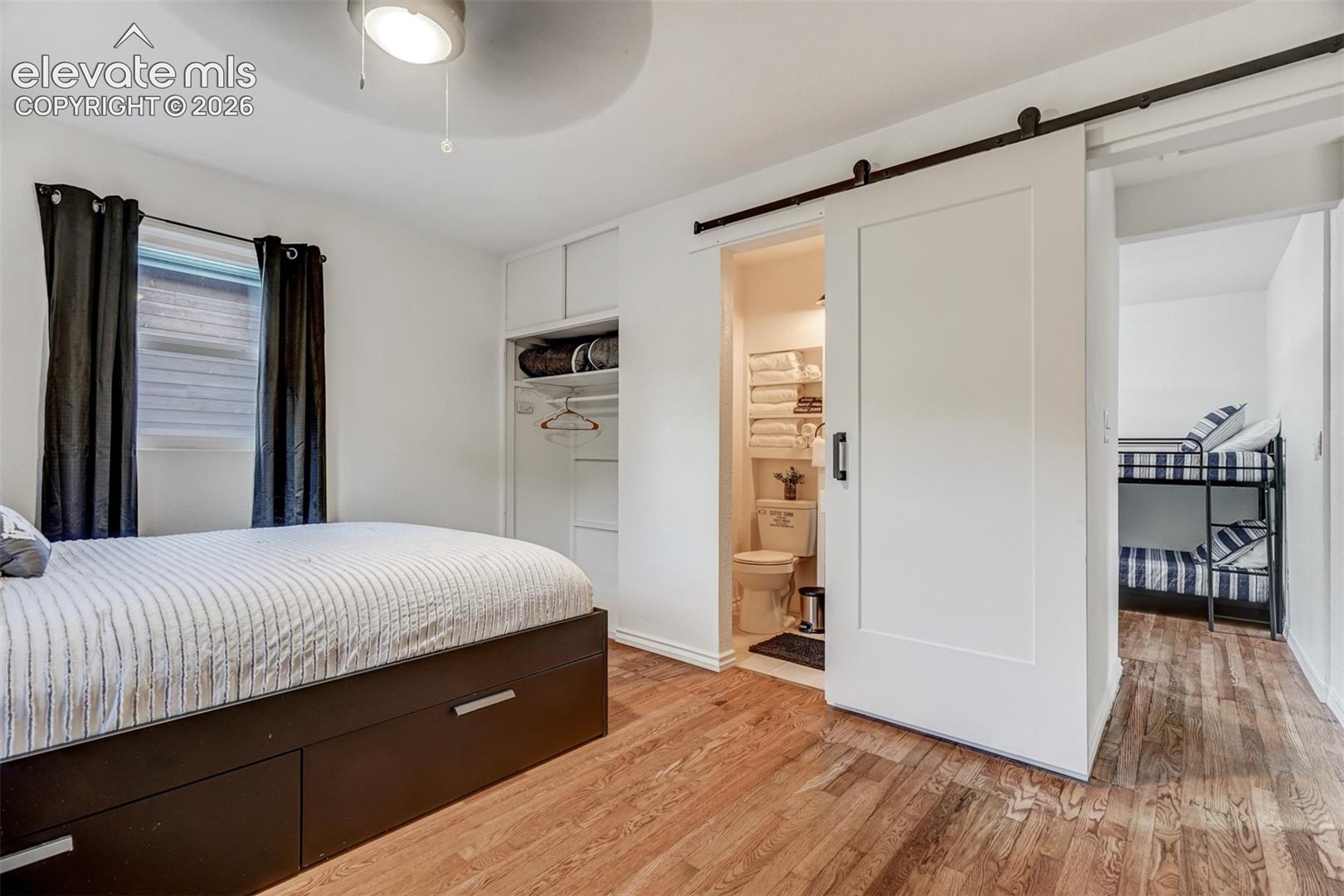 Wood-finish flooring, built-in storage, modern ceiling fan, white barn door, and a full bathroom with toilet