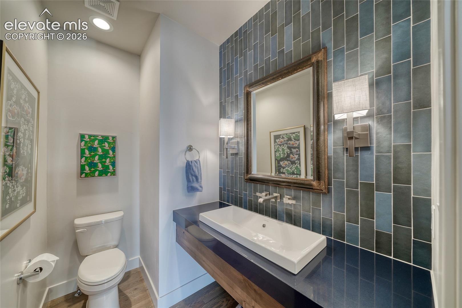 Floating vanity featuring a dark countertop, integrated rectangular vessel sink, wall-mounted faucet, floor-to-ceiling vertical blue tile backsplash, and dual wall-mounted light fixtures