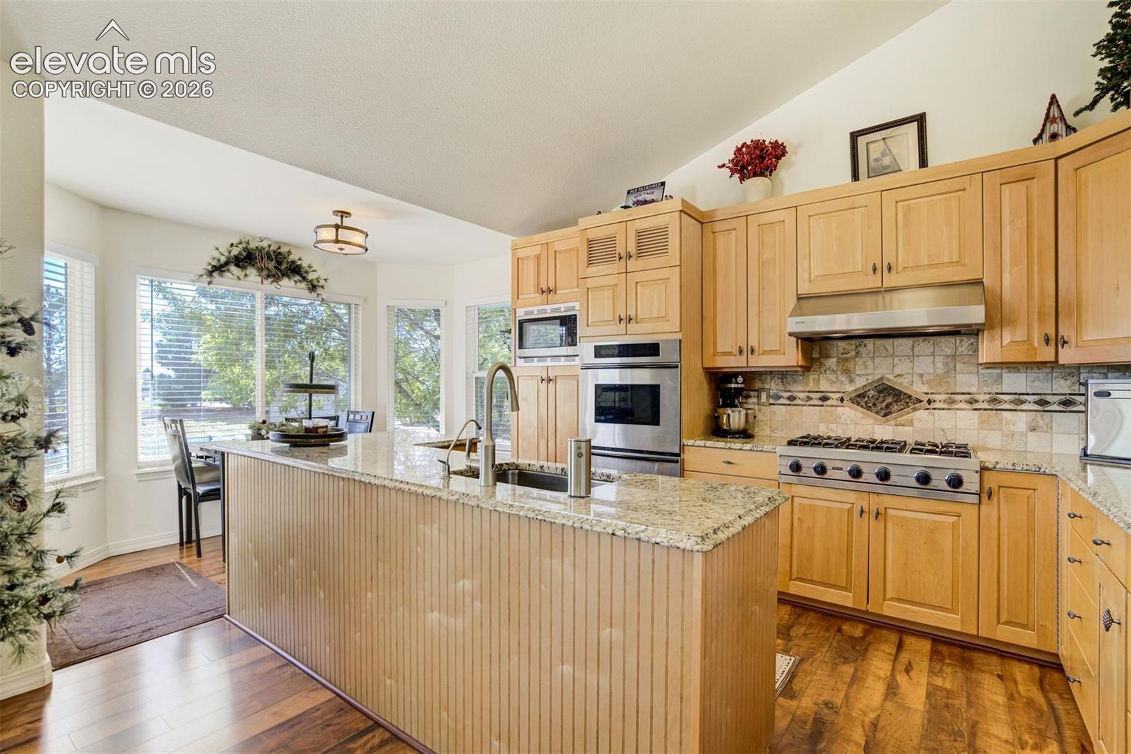 Kitchen featuring light-tone wood cabinetry, granite countertops, stainless steel wall oven and gas cooktop, tile backsplash, and wood-finish flooring