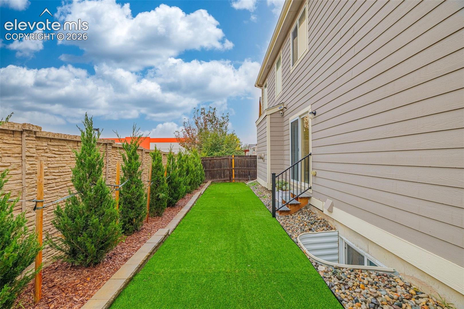 Side yard featuring synthetic turf, structured evergreen plantings, decorative stone ground cover, a textured privacy wall, and an egress window well