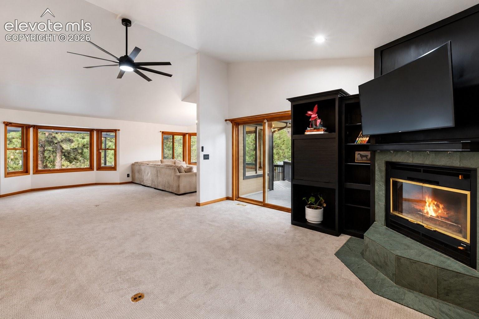 Vaulted ceilings with a multi-blade ceiling fan, a floor-to-ceiling stone-surround fireplace, built-in cabinetry, large bay windows, and sliding glass doors