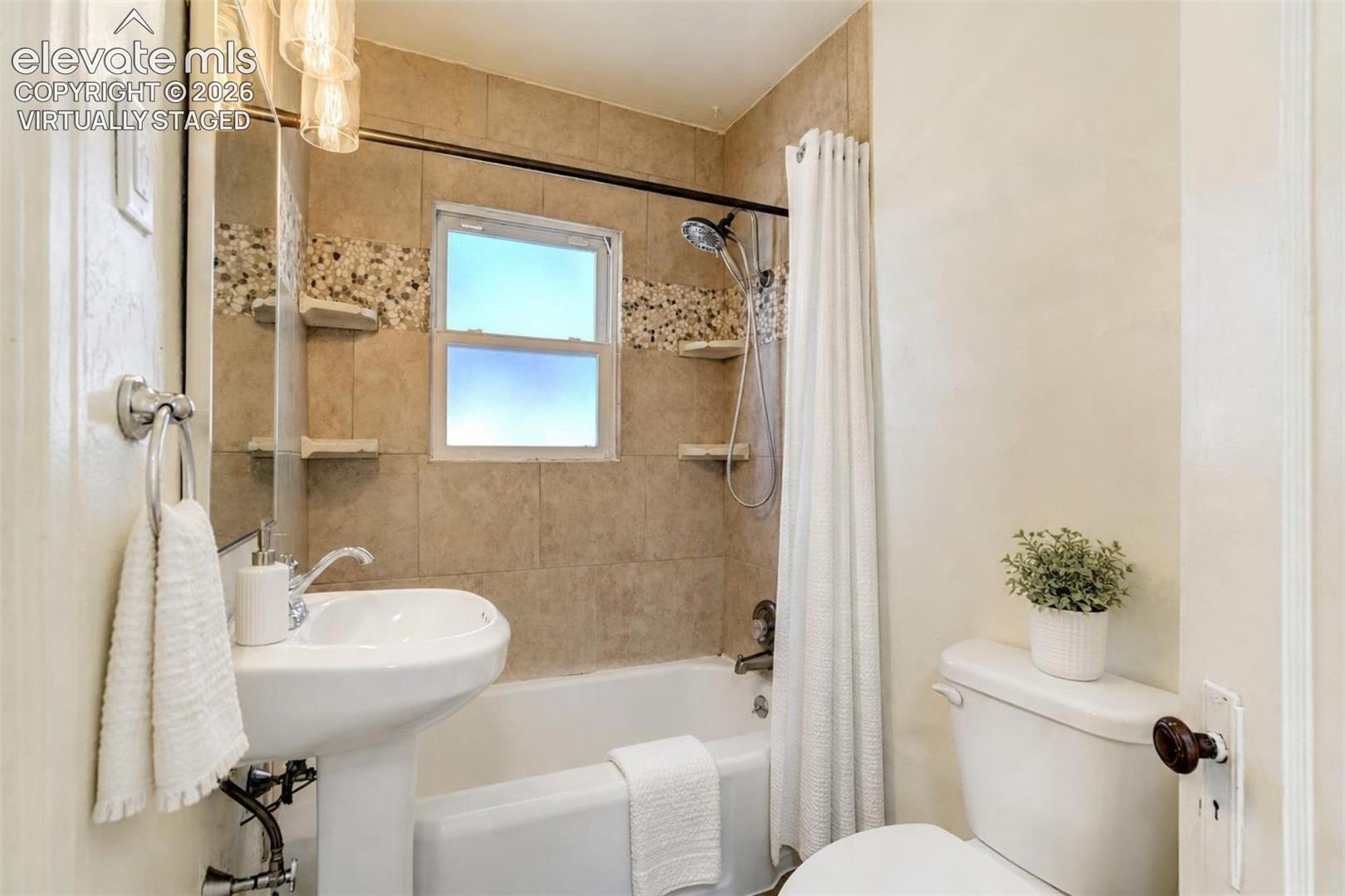 Virtually Staged.  Bathroom featuring a white pedestal sink, tiled shower surround with decorative stone inlay, built-in corner shelving, handheld shower fixture, and a large wall-mounted mirror