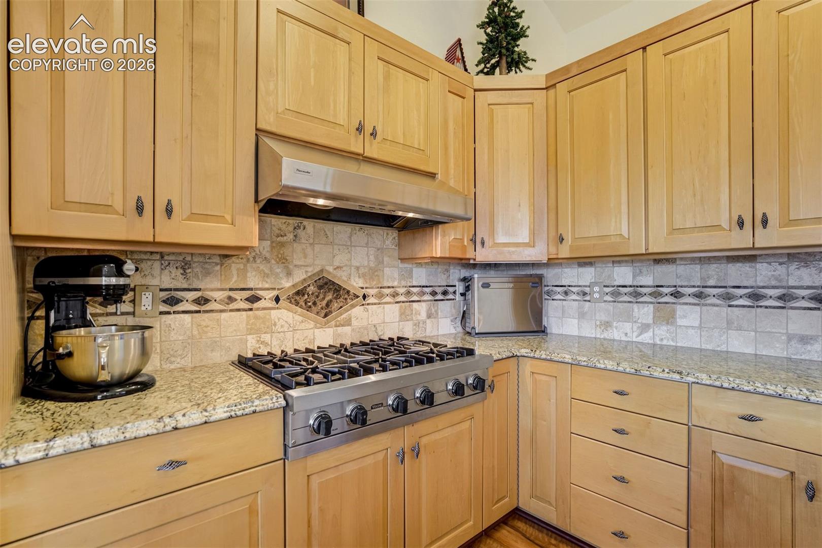 Light wood cabinetry with granite countertops, tiled backsplash with decorative inlay, stainless steel gas range, and matching stainless steel range hood
