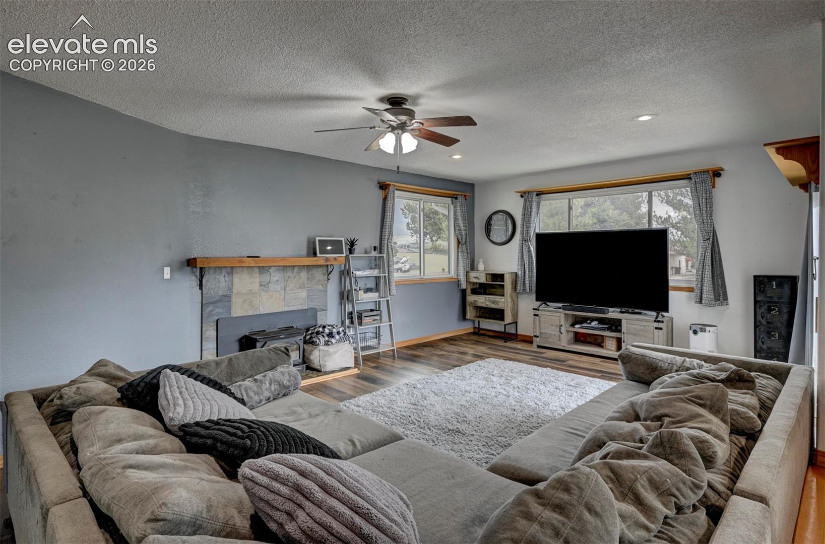 Living area featuring wood-finish flooring, a ceiling fan, and a stone-surround fireplace with a wood mantle