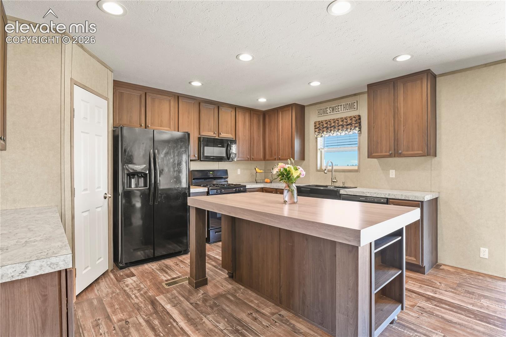 Kitchen featuring wood-finish cabinetry, stone-look laminate countertops, built-in black appliances, recessed lighting, and a central wood-finish island with integrated shelving