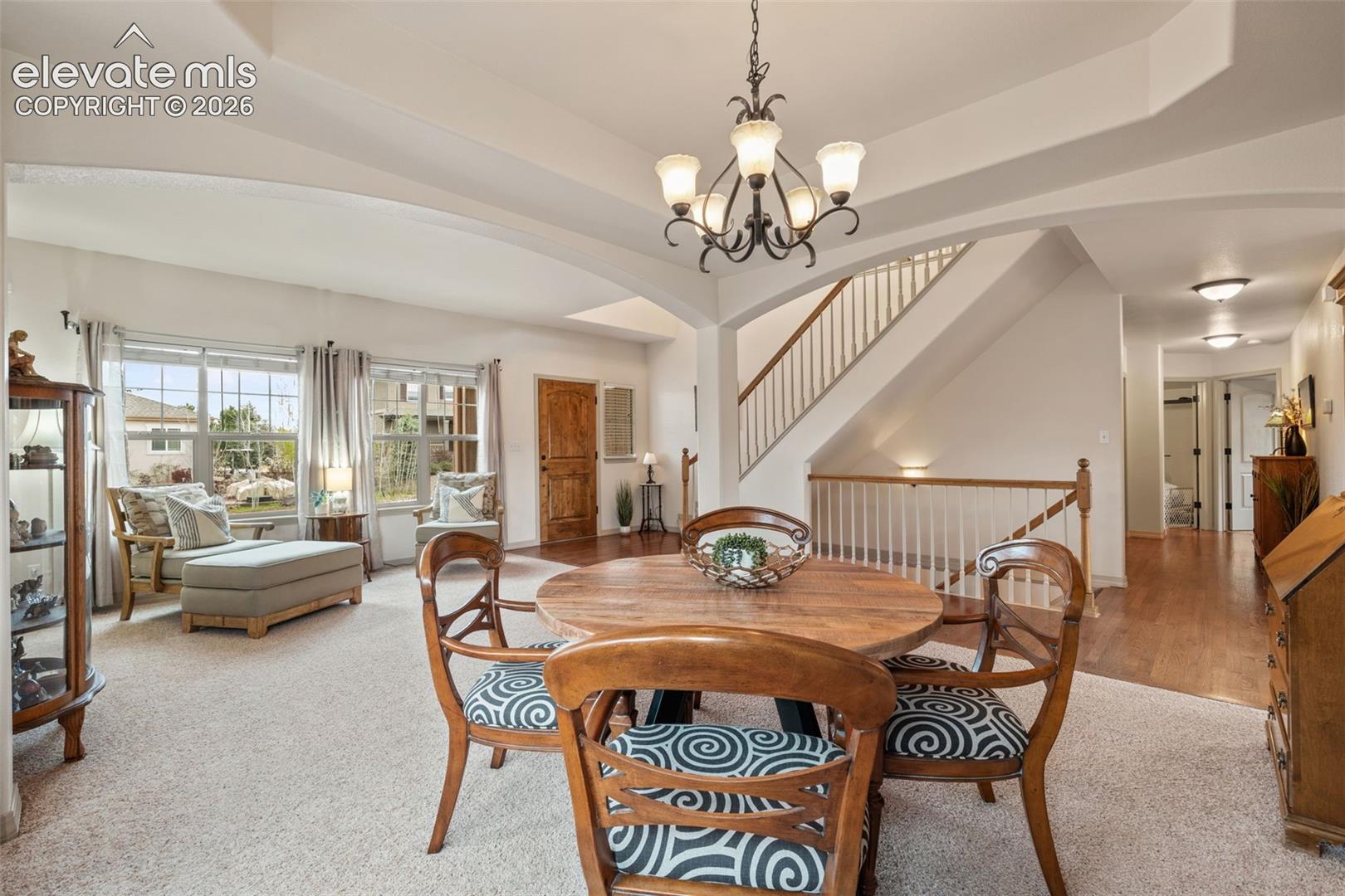 Open-concept living area featuring a formal dining space with an overhead chandelier, a carpeted seating area with large windows, a wooden front door, and a staircase with a wood handrail and white balusters
