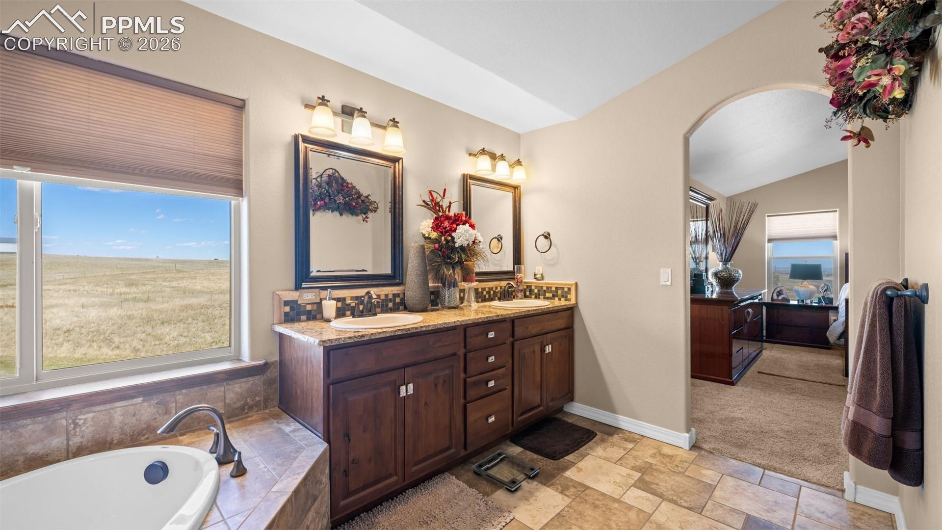 Ensuite bathroom featuring double vanity, a garden tub, light stone finish flooring, plenty of natural light, and lofted ceiling