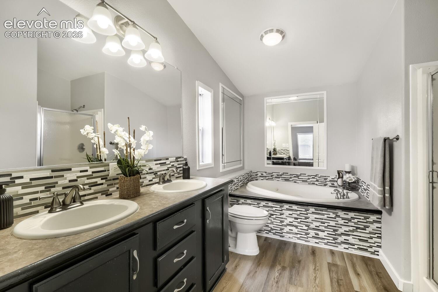 Bathroom featuring a dual-sink vanity with dark cabinetry, integrated stone-finish countertop, decorative linear mosaic tile backsplash, soaking tub with matching tile surround, and wood-finish flooring