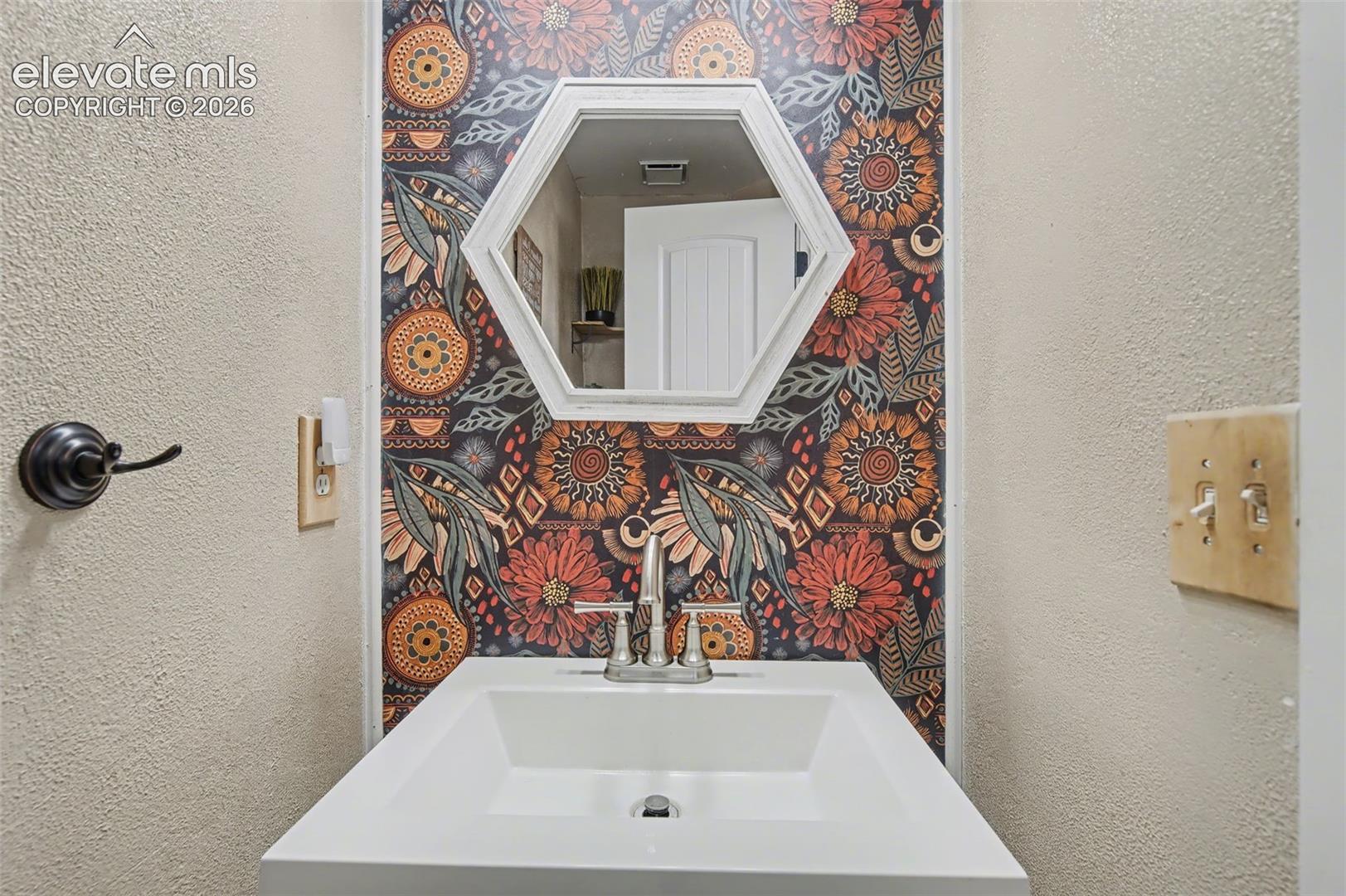 Wall-mounted hexagonal mirror with white frame, patterned wall covering, and white pedestal sink featuring a dual-handle metal faucet