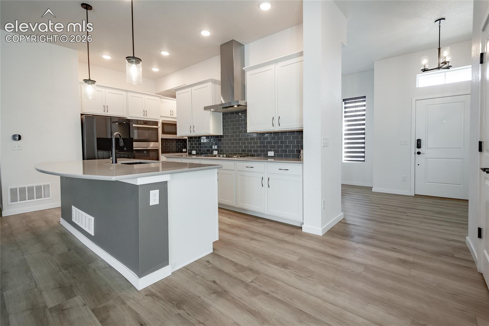 Kitchen featuring white cabinetry, wood-finish flooring, a large center island with quartz countertop, a stainless steel range hood, and a dark-toned tile backsplash