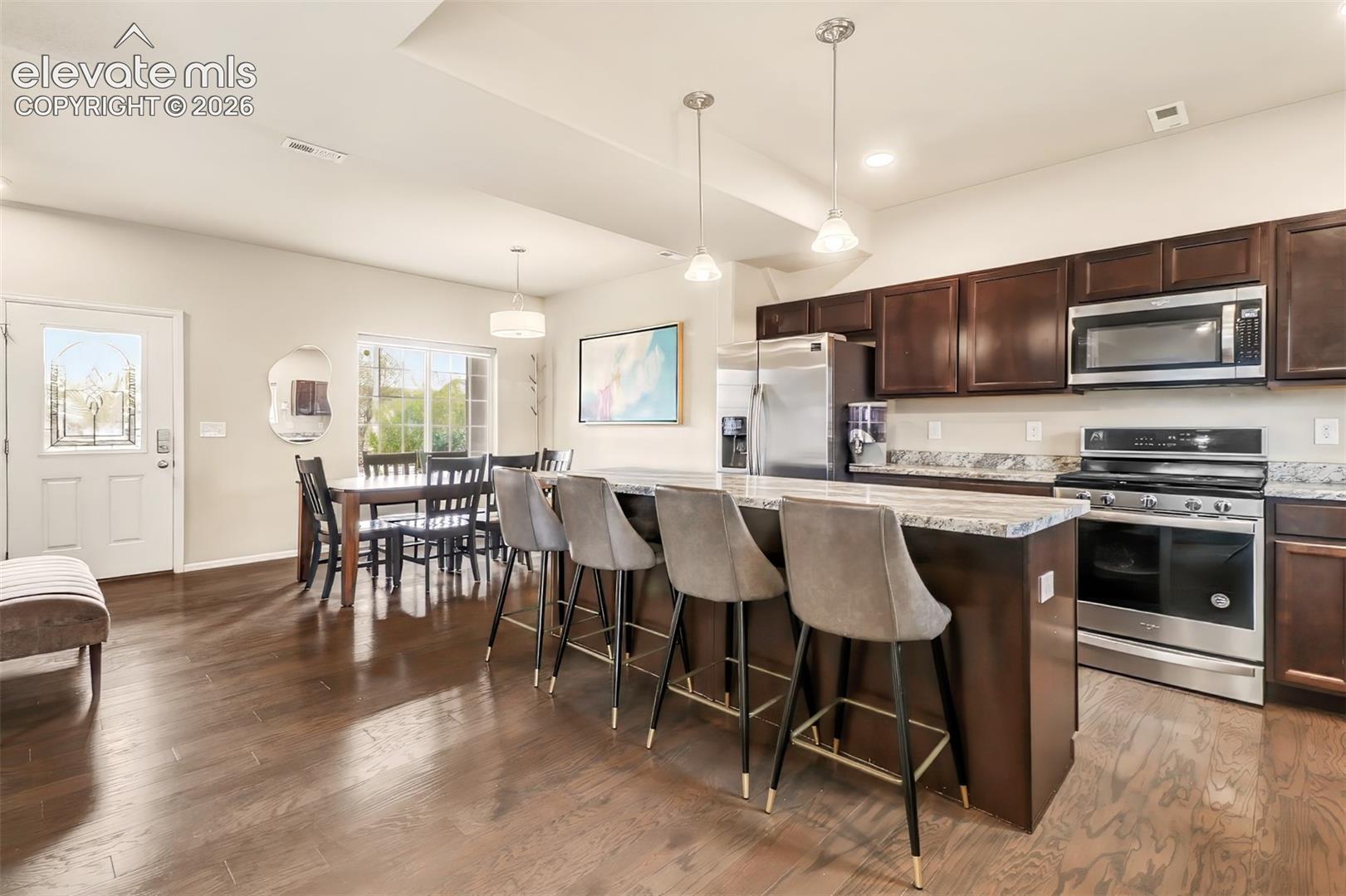 Kitchen featuring dark wood cabinetry, stone-patterned countertops, stainless steel appliances, a center island with pendant lighting, and wood-finish flooring