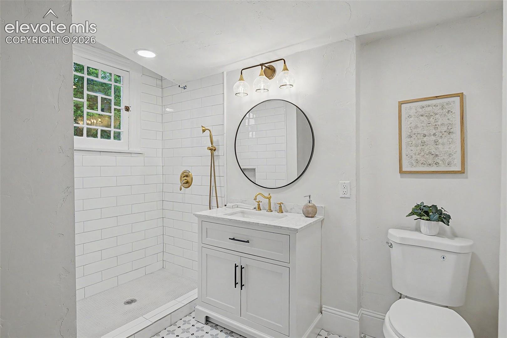 Contemporary bathroom featuring a subway-tiled shower with gold-finish fixtures, a white vanity with a marble-finish countertop, a circular mirror, and patterned floor tiling