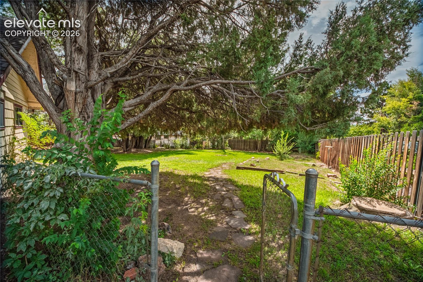 Expansive yard space featuring a chain-link fence and gate, a stone pathway, a wood privacy fence, and mature trees