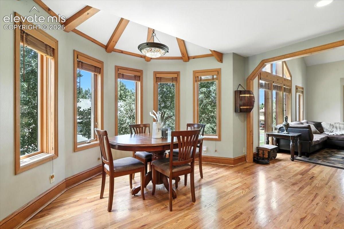 Polygonal dining area featuring wood-finish flooring, vaulted ceiling with exposed wood beams, natural wood window trim, and a multi-light flush-mount chandelier