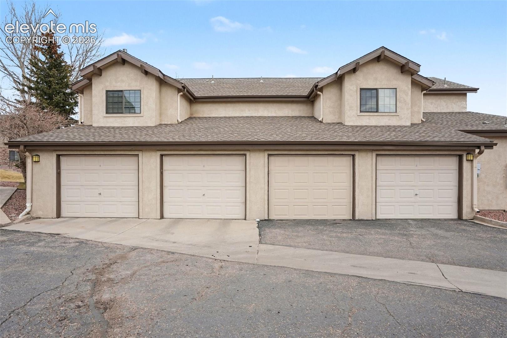 Stucco exterior featuring four garage bays, dual gabled rooflines, asphalt shingle roofing, and exterior wall-mounted lighting