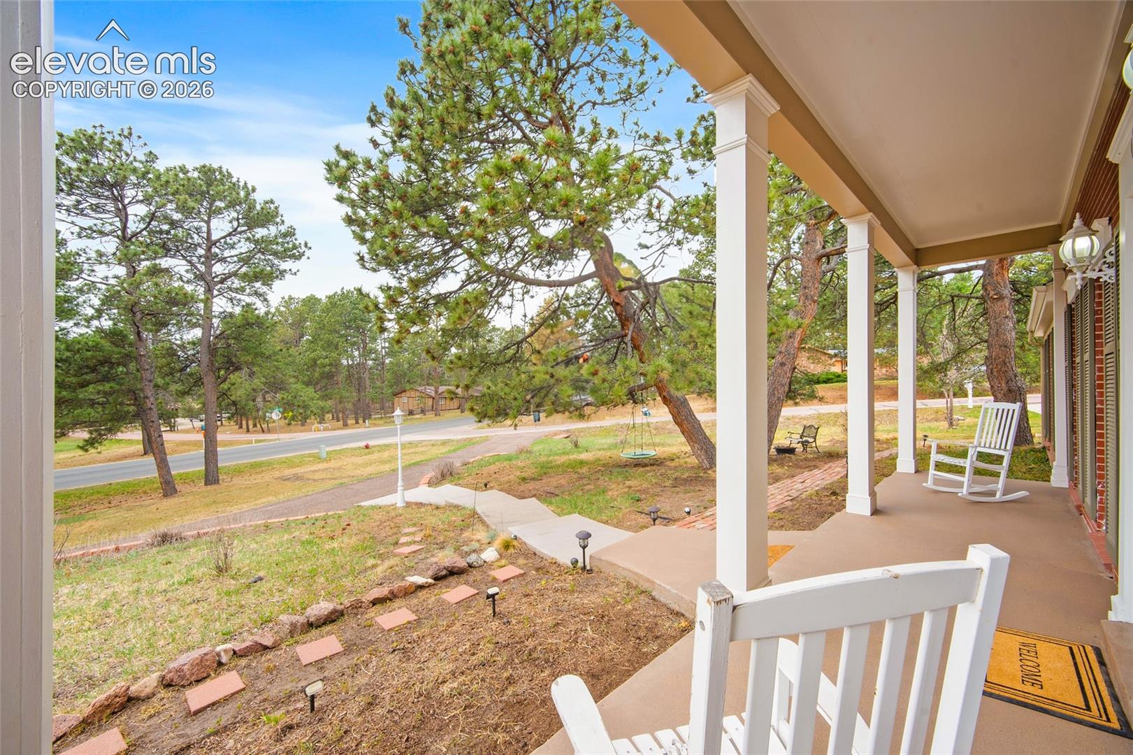 Covered front porch featuring white structural columns, concrete walkway, brick landscape borders, and mature pine trees