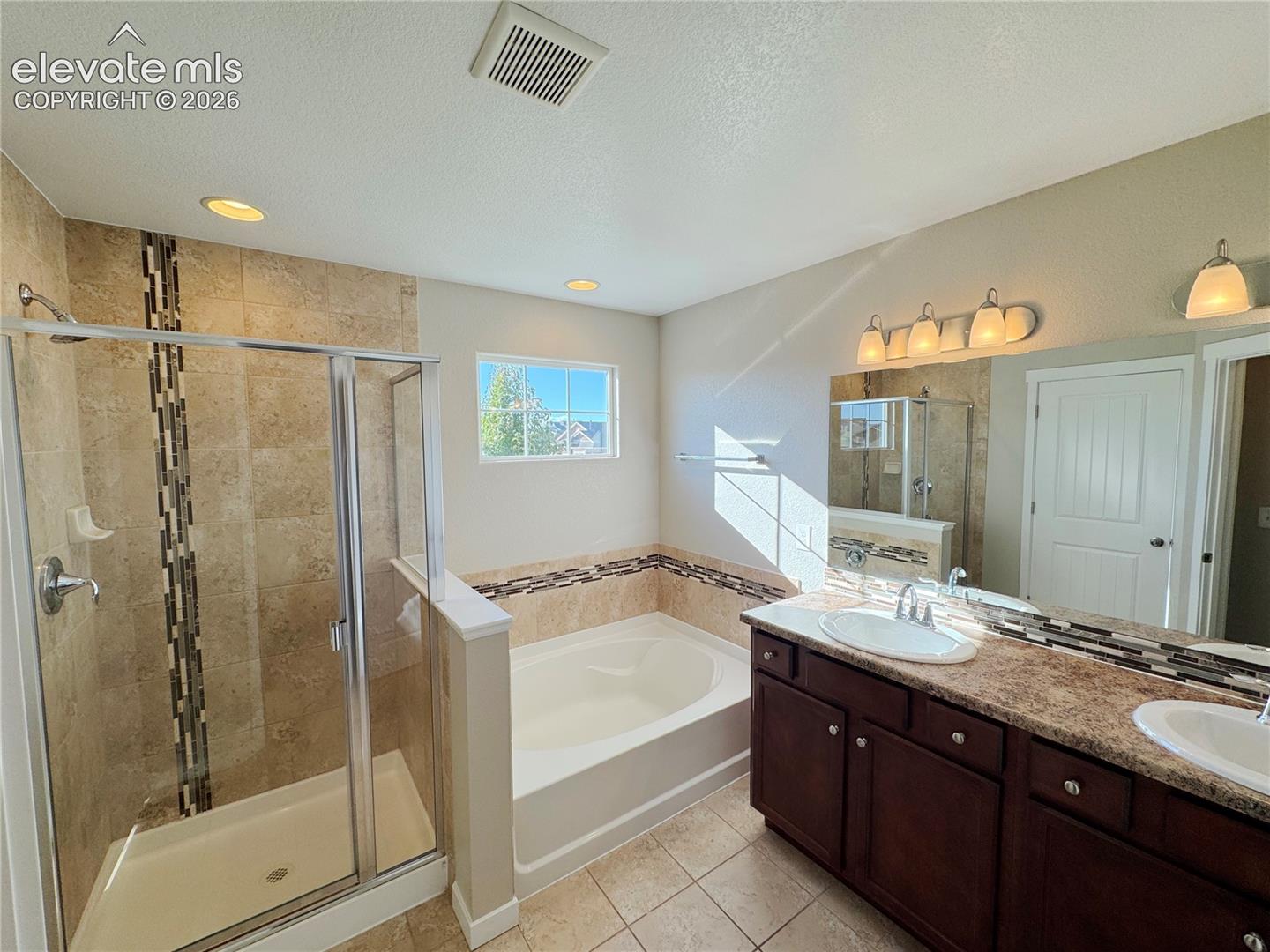 Bathroom featuring a dual-sink vanity with dark-finish cabinetry, a built-in soaking tub with decorative tile inlay, a separate shower enclosure with stone-style wall tile, and recessed lighting