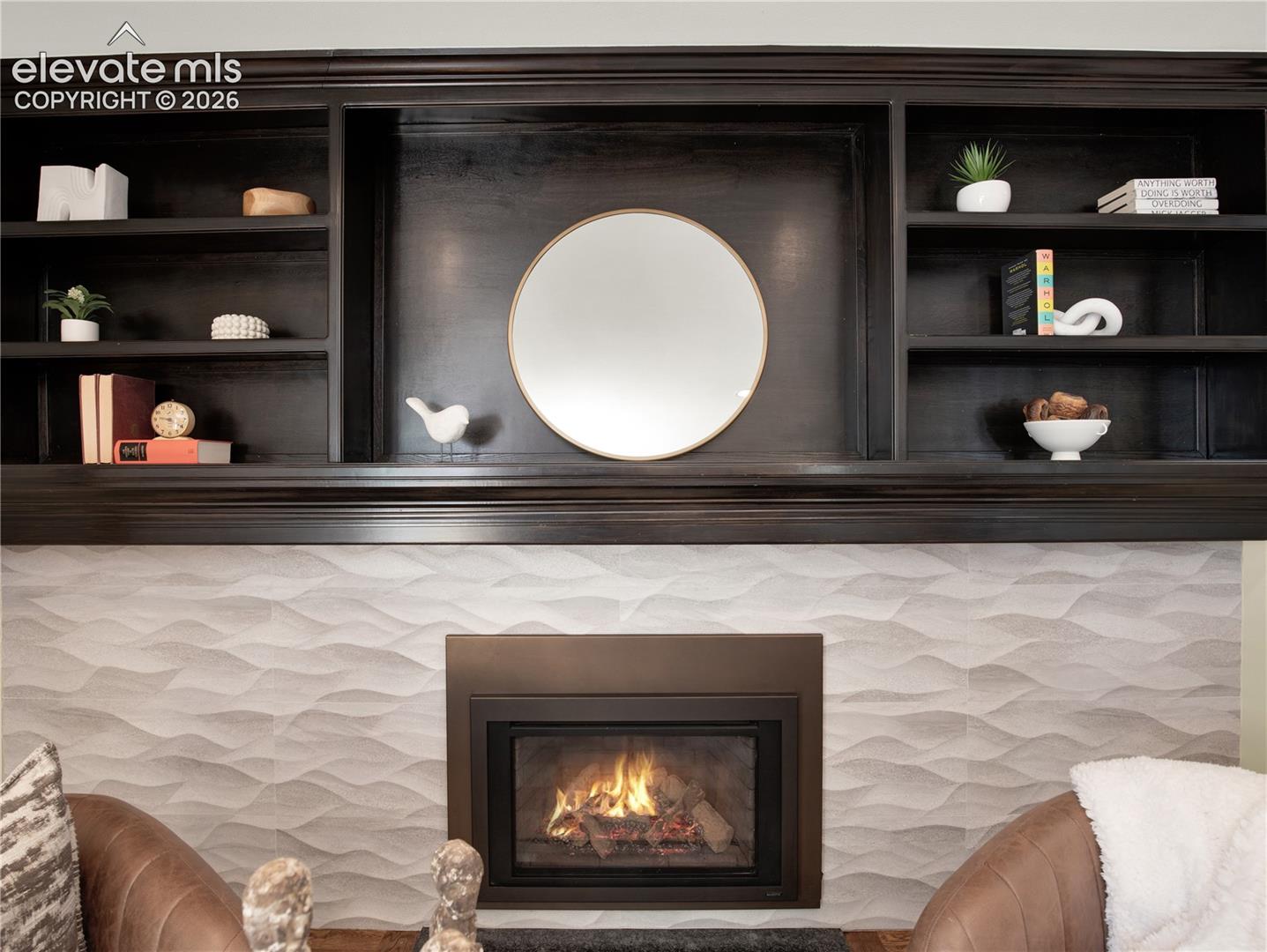 Built-in dark-finish cabinetry featuring open shelving, a central circular mirror, a textured light-toned tile surround, and a rectangular metal fireplace insert