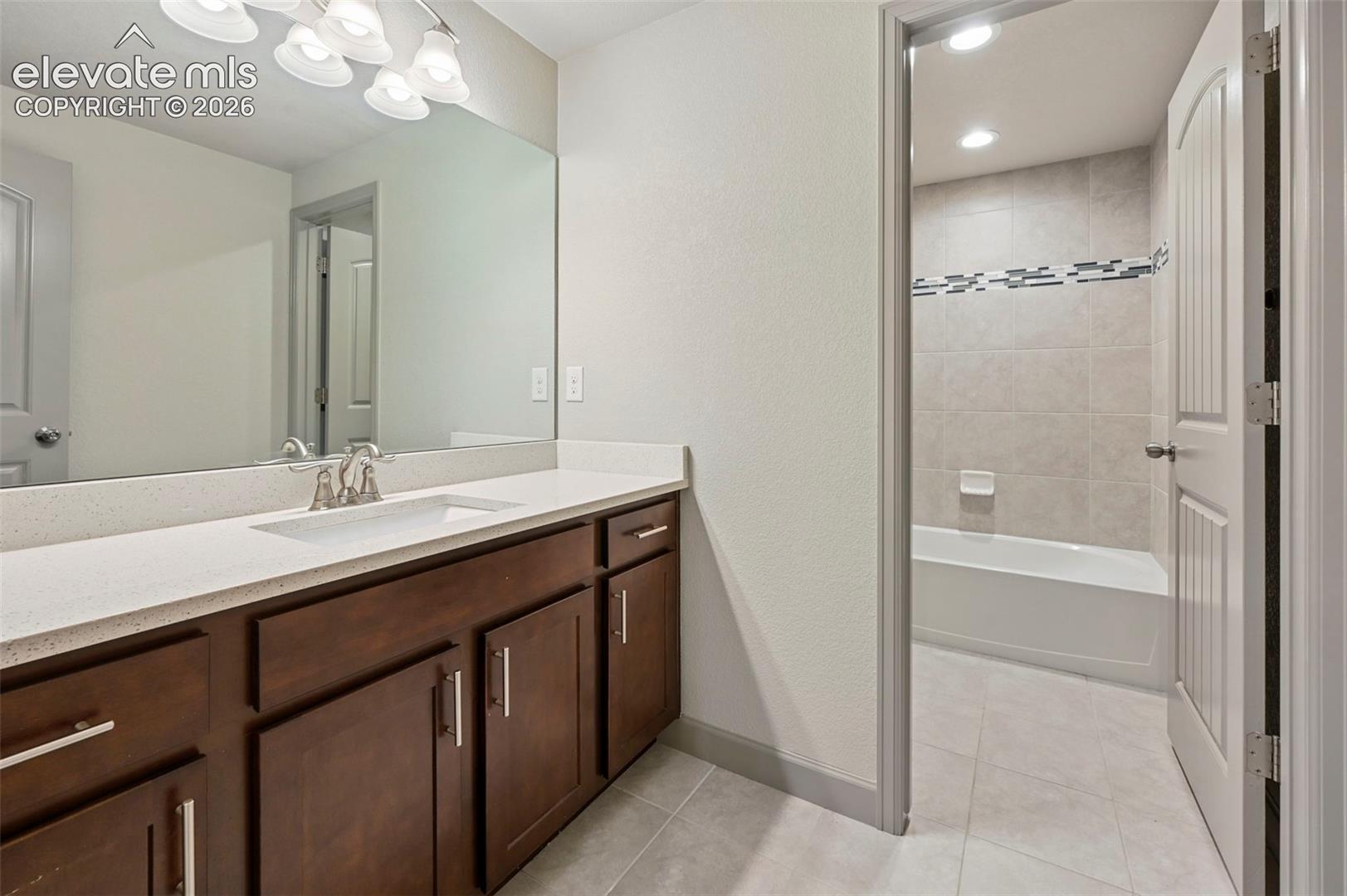 Bathroom featuring a dark wood-finish vanity with a light-toned solid surface countertop, an undermount rectangular sink, a full-width wall mirror, and a tiled tub-shower combination with decorative accent tile