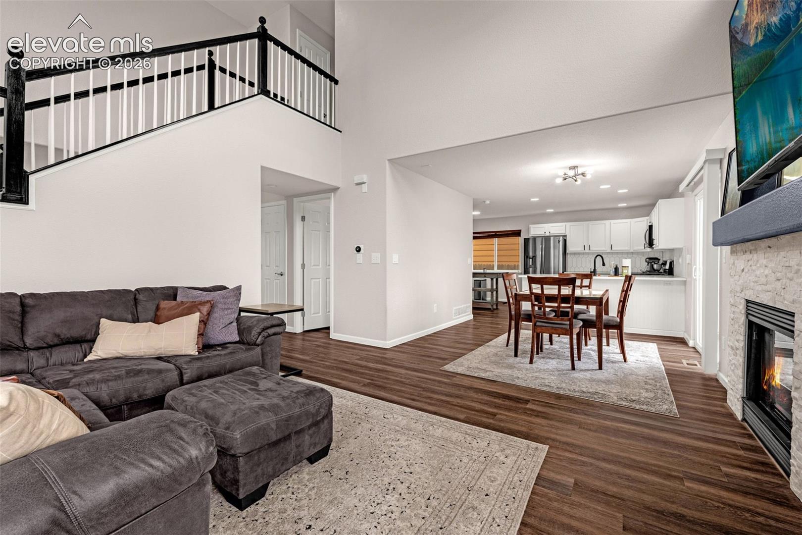 Open-concept interior featuring wood-finish flooring, a two-story ceiling, a staircase with a black and white railing, and a stone-surround fireplace