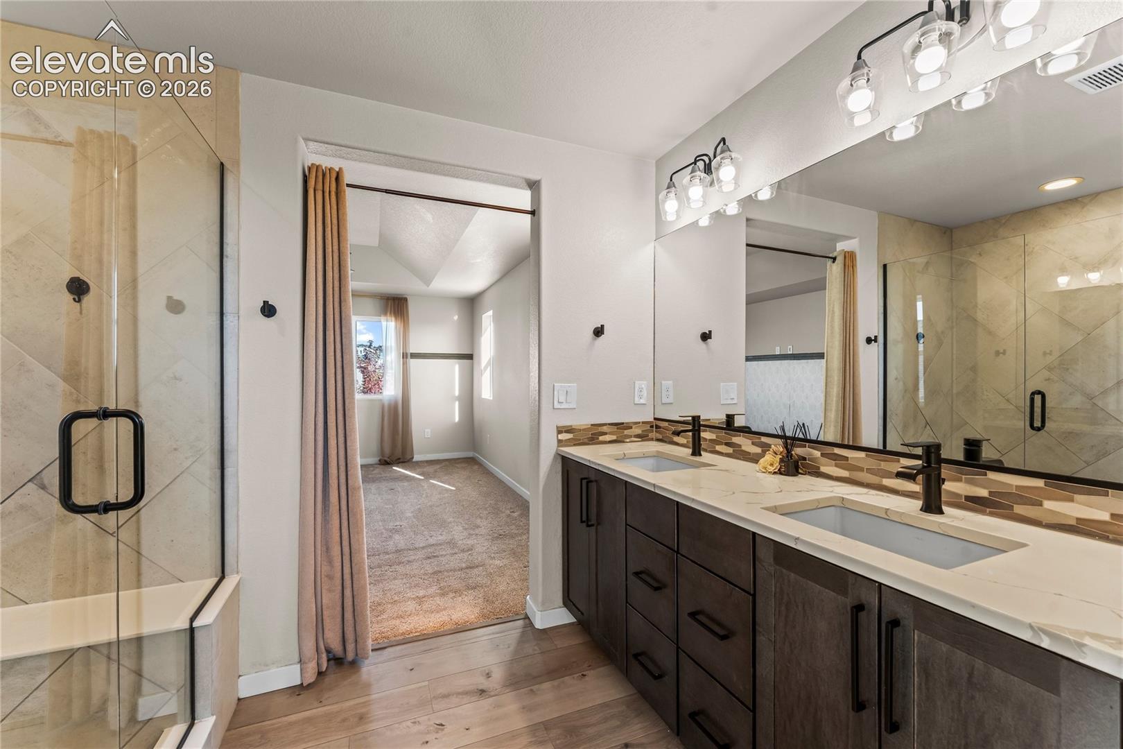 Dual-sink vanity with stone-finish countertop, dark wood-finish cabinetry, frameless glass shower enclosure with neutral-toned tile, and large wall-mounted mirror
