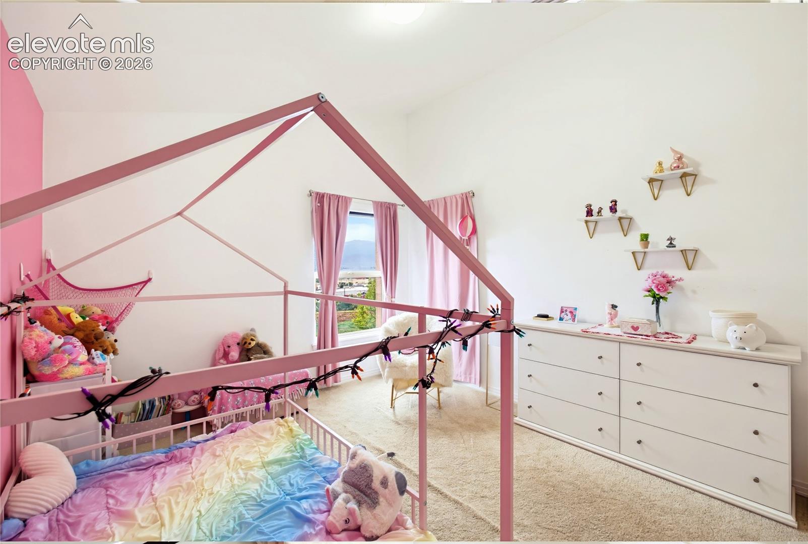Vaulted ceiling, white walls, carpet flooring, window with light pink drapes, and built-in floating shelves