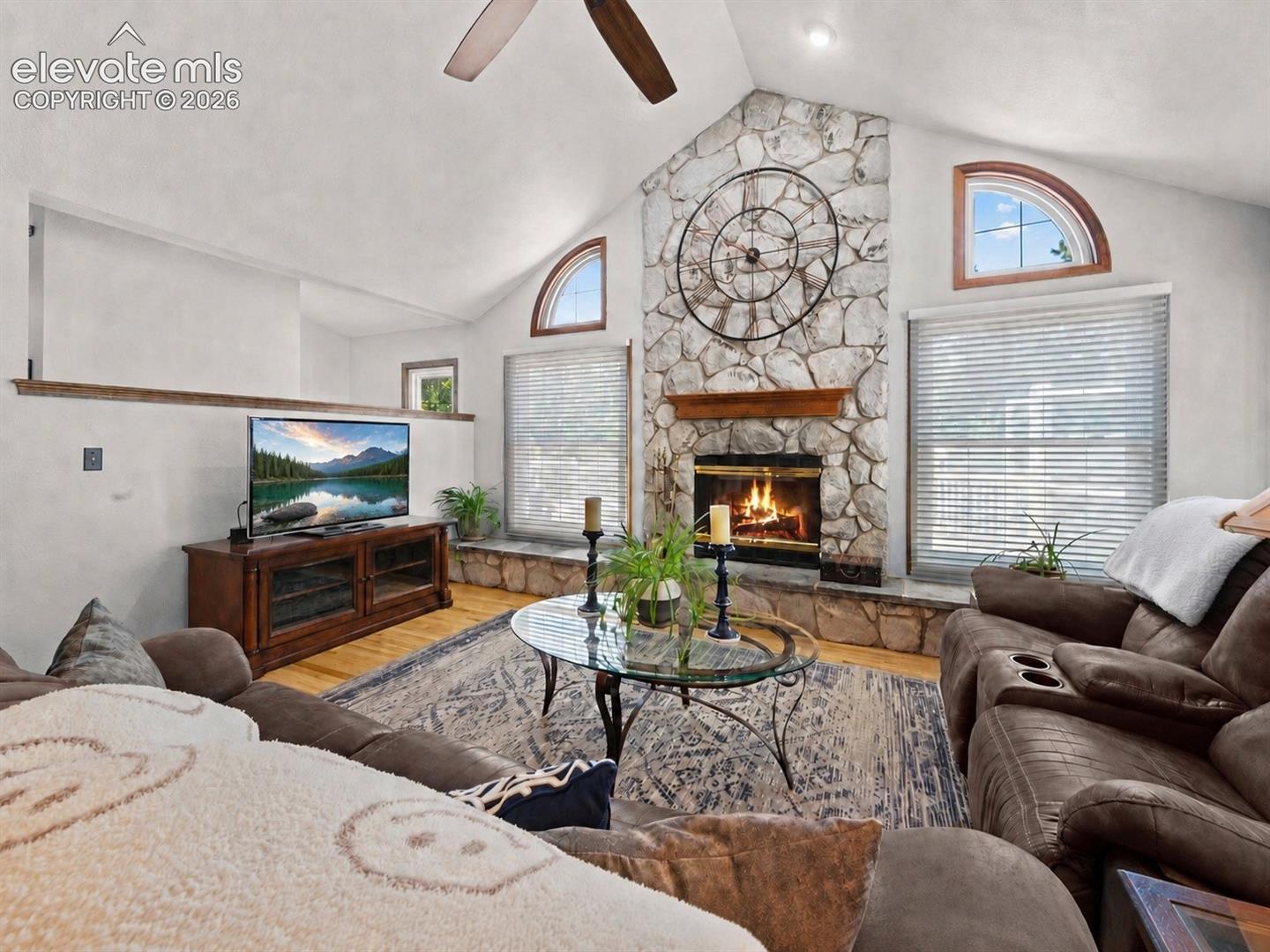 Vaulted ceiling with a ceiling fan, stone-clad fireplace with a wood mantle, arched windows, and wood-finish flooring