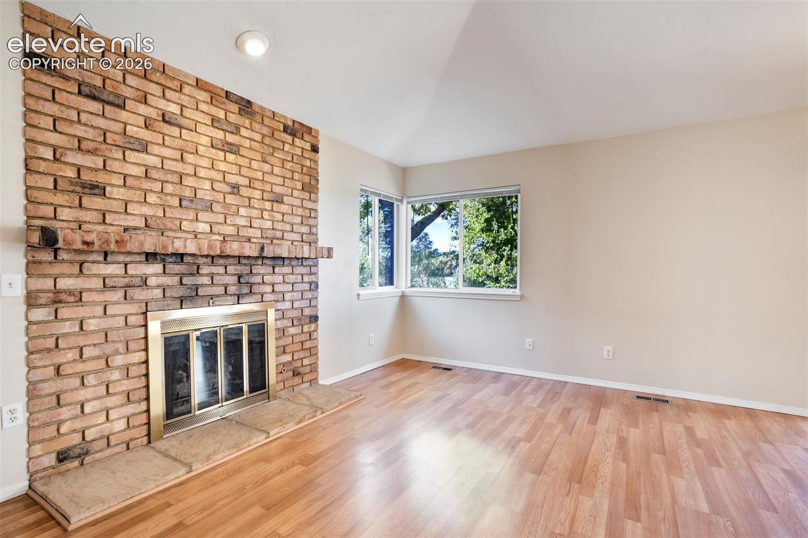 Floor-to-ceiling brick fireplace with a wood-finish mantel, stone hearth, brass-framed firebox, wood-finish flooring, and corner windows