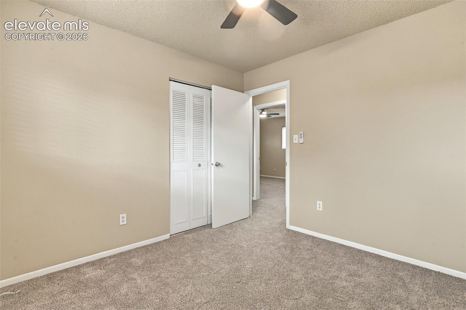Neutral-toned room featuring textured ceiling, carpeting, and louvered closet doors with a ceiling-mounted fan