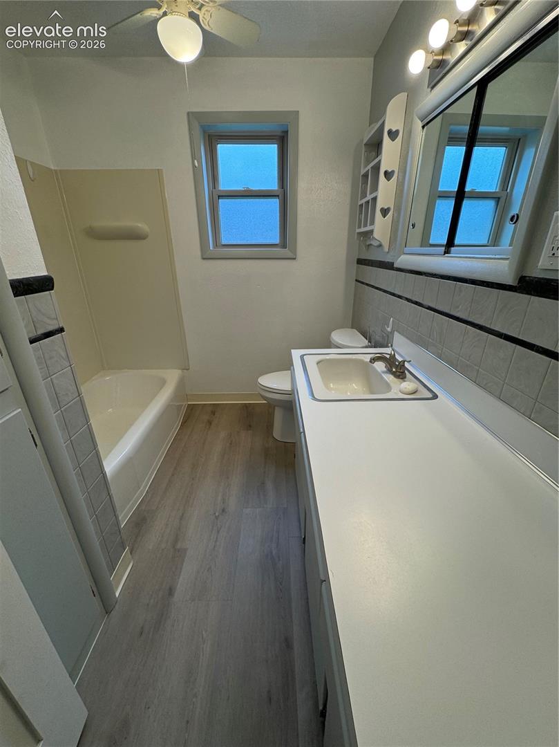 Bathroom featuring wood-finish flooring, a white vanity with an integrated sink, a tiled tub-shower surround, and a ceiling-mounted light fixture with a fan