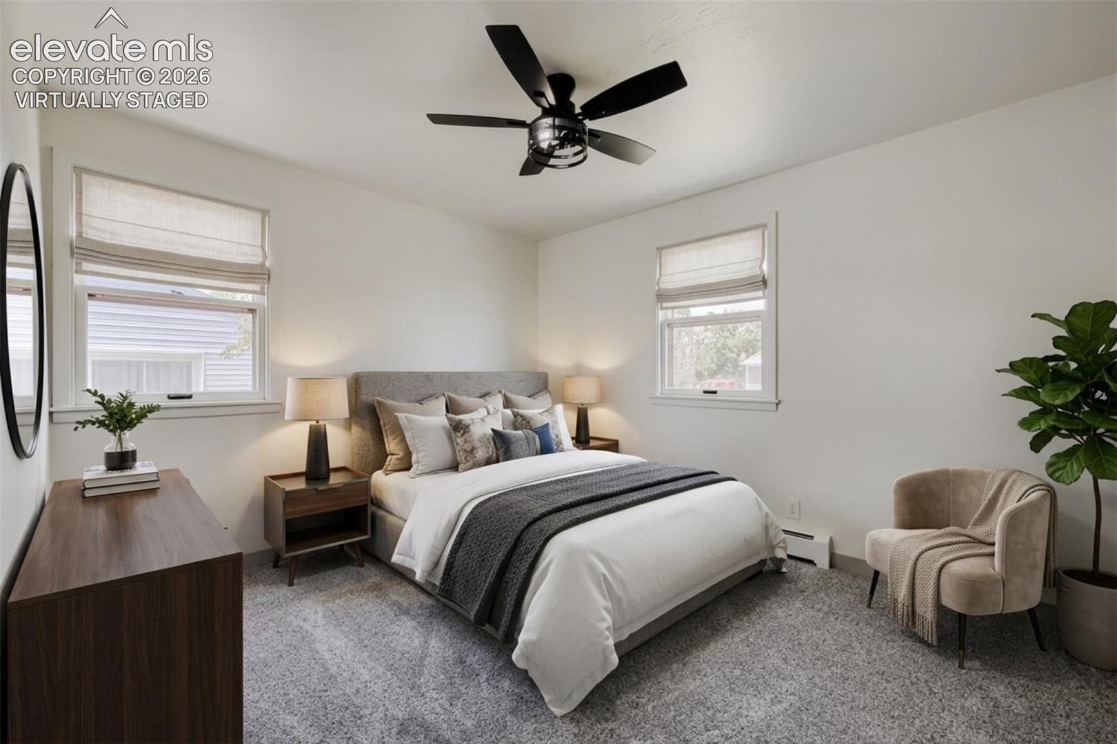 Recessed lighting, ceiling fan with matte black finish, light gray upholstered headboard, two windows with blinds, and light gray carpeting. Virtually staged.