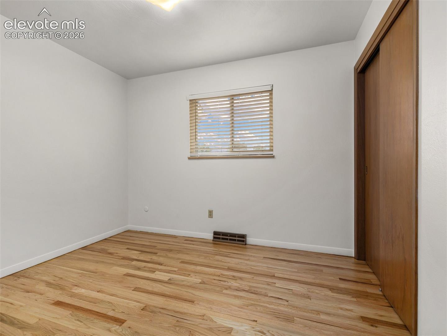 Hardwood flooring, natural light through window, horizontal blinds, white walls, and built-in closet with wood-finish sliding doors