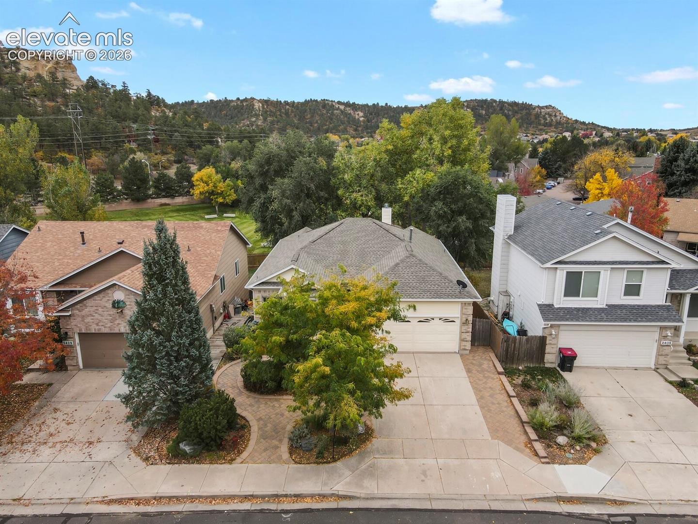 Single-story residential exterior featuring an attached two-car garage, stone facade accents, shingled roofing, and a paved driveway, set against a mountainous landscape