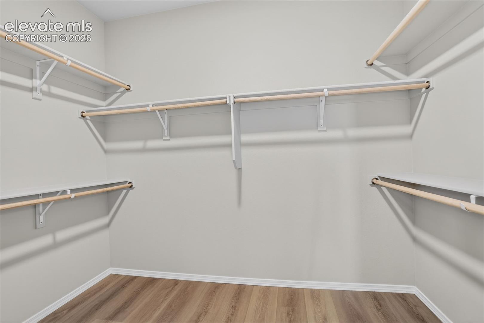 Expansive closet space featuring wood-finish flooring, light gray wall paint, white baseboards, and built-in white shelving with natural wood rods
