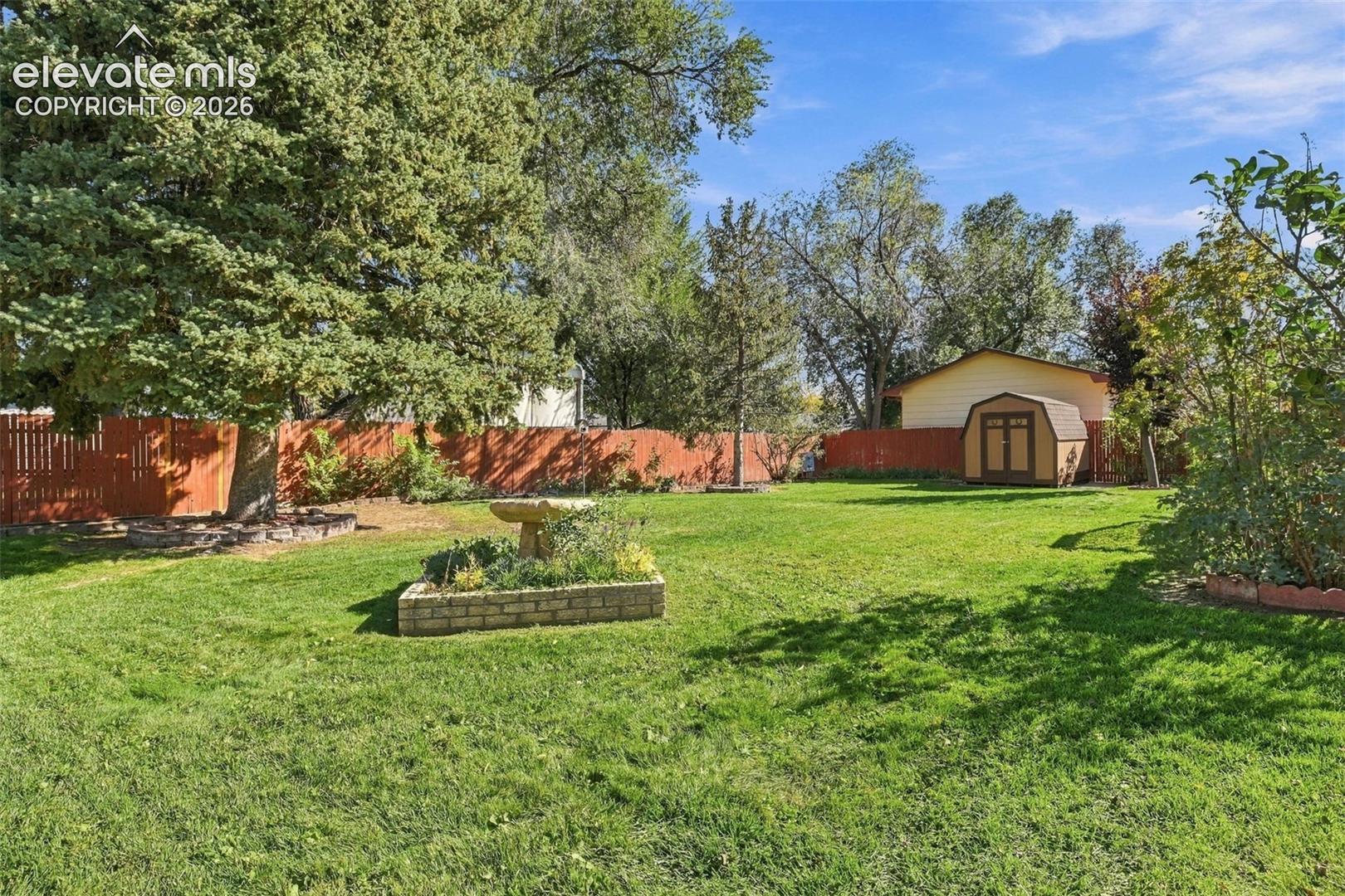 Expansive backyard featuring a mature canopy of trees, a wood-slat privacy fence, a brick-enclosed garden bed with a stone fountain, and a detached storage structure
