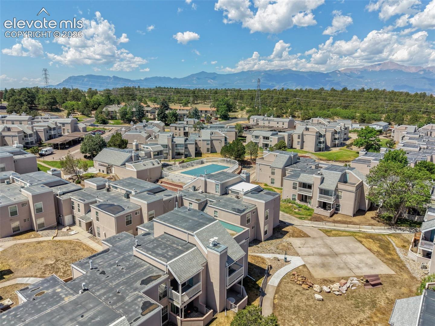 Aerial perspective of a multi-unit residential community featuring a central outdoor swimming pool, concrete walkways, and mountain range backdrop
