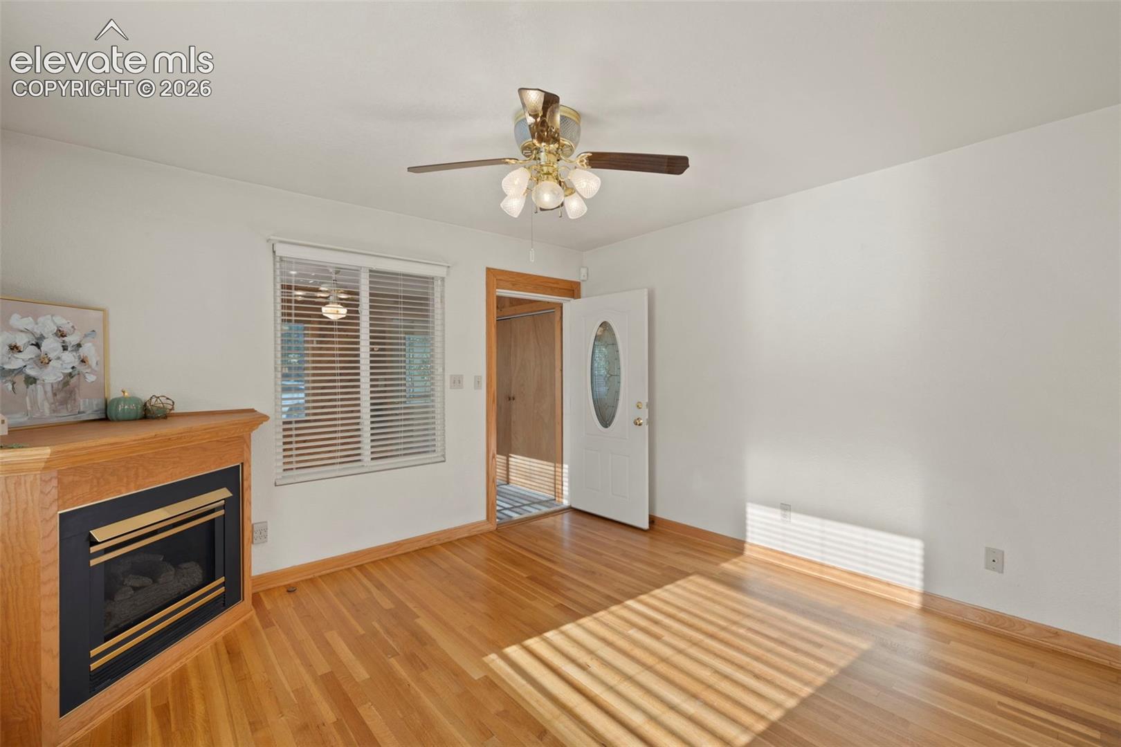 Wood-finish flooring, built-in fireplace with wood mantel, white walls, ceiling fan with integrated lighting, and white entry door with decorative glass inlay