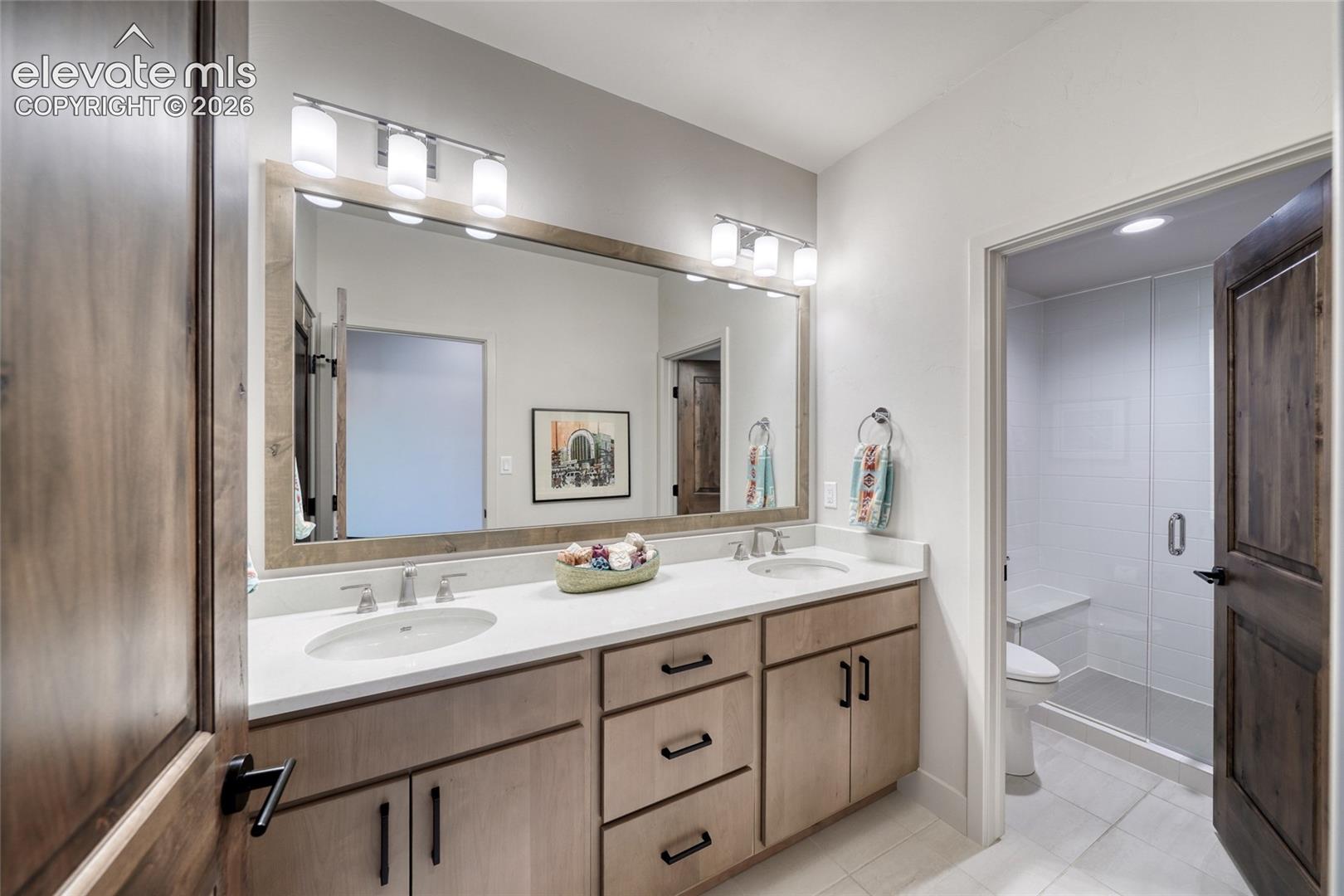 Dual-sink vanity featuring a solid surface countertop, light wood cabinetry with matte black hardware, large-scale framed mirror, and two multi-bulb vanity lights