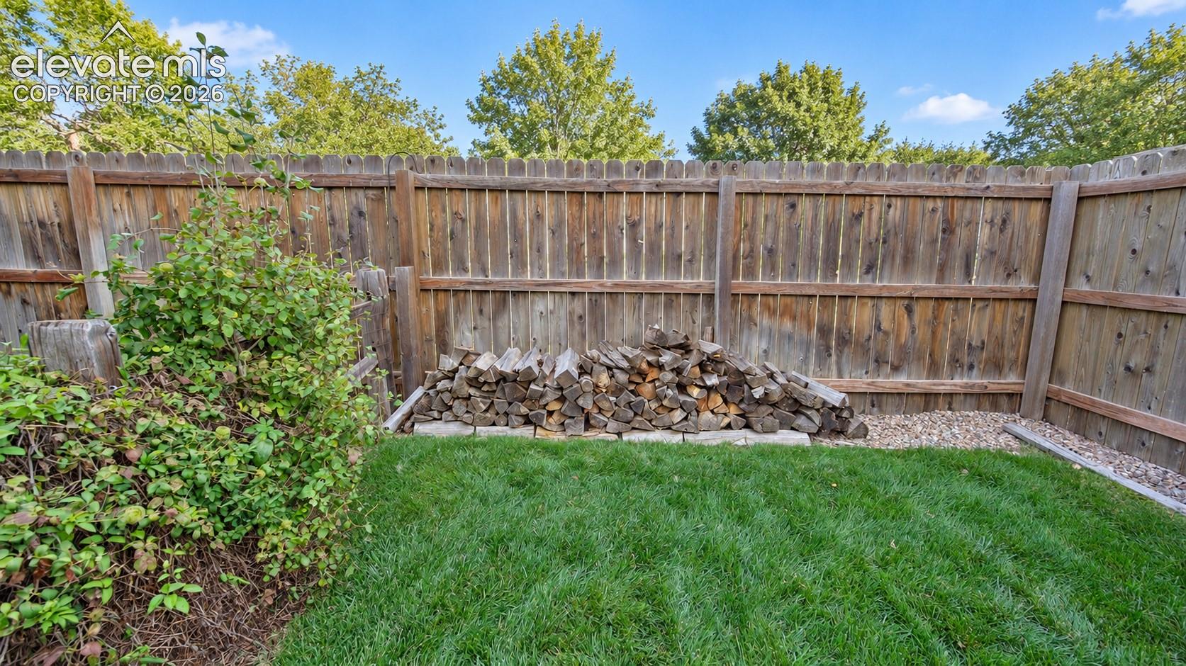 Fenced yard with lush landscaping, mature trees, green lawn, stack of firewood for the internal Family Room fireplace.