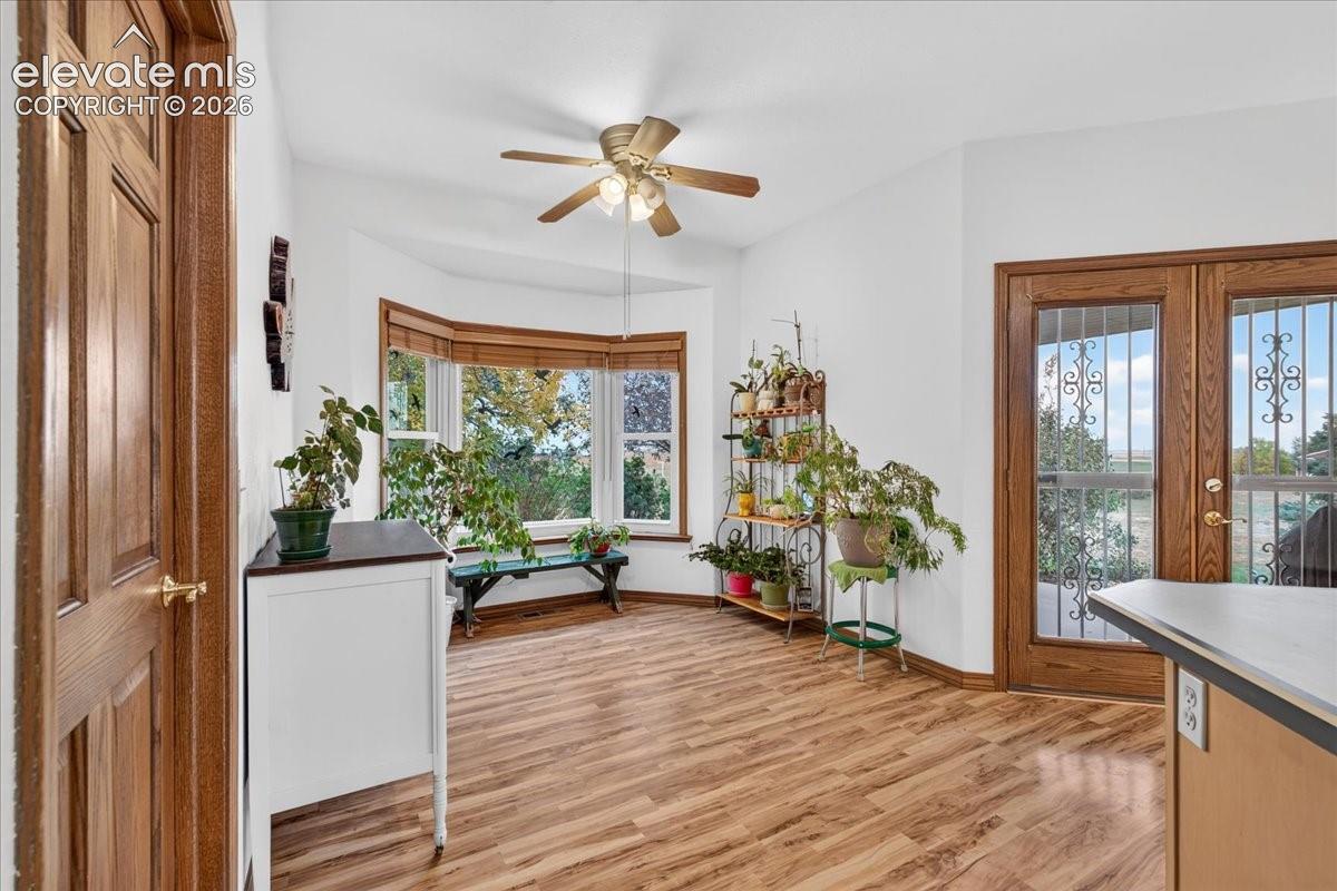 Bay window with natural light, wood-finish flooring, multi-pane glass door with decorative metal security grating, and a multi-blade ceiling fan with integrated lighting