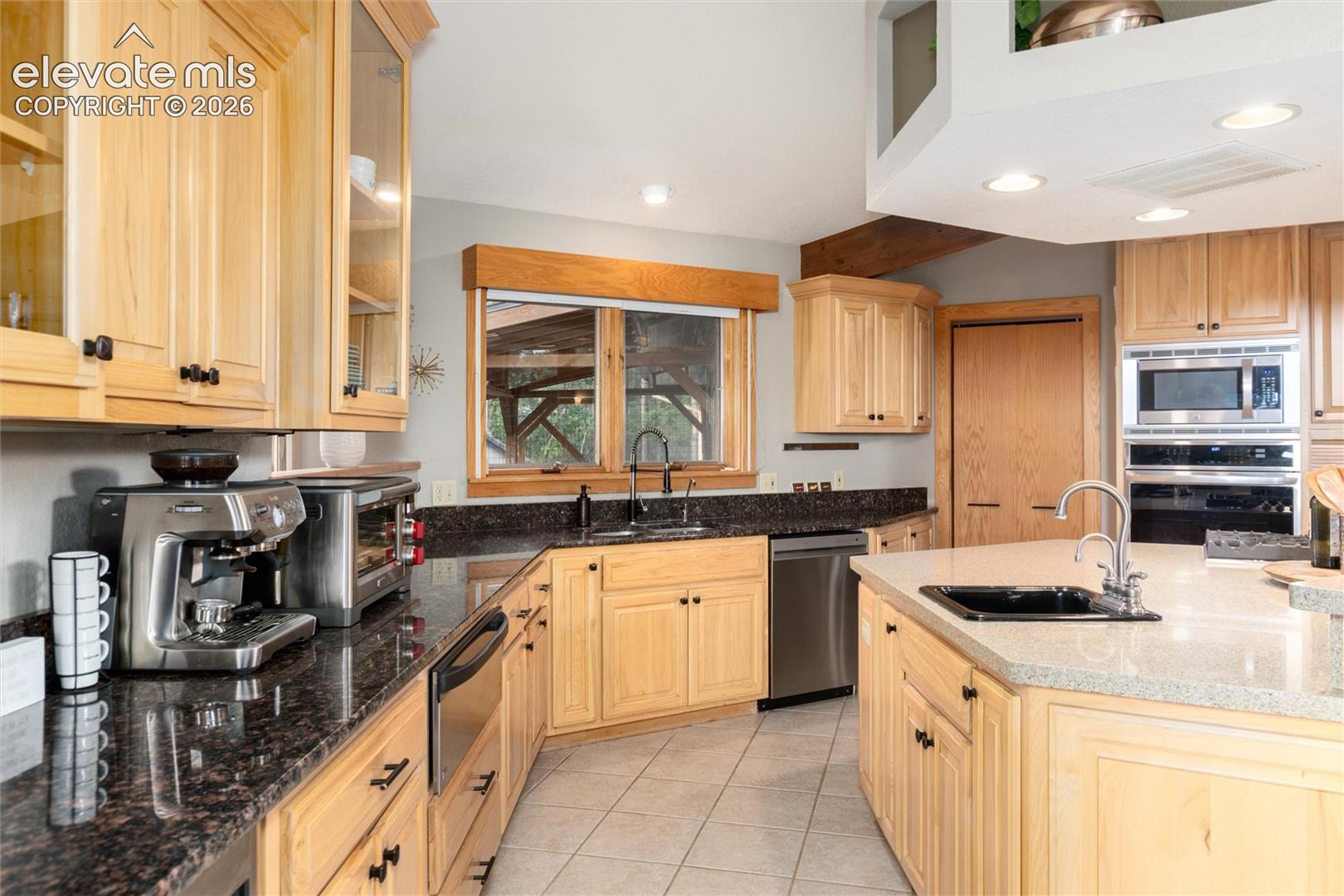 Kitchen featuring light wood cabinetry, dark stone countertops, stainless steel appliances, a center island with sink and hot water tap, and recessed lighting