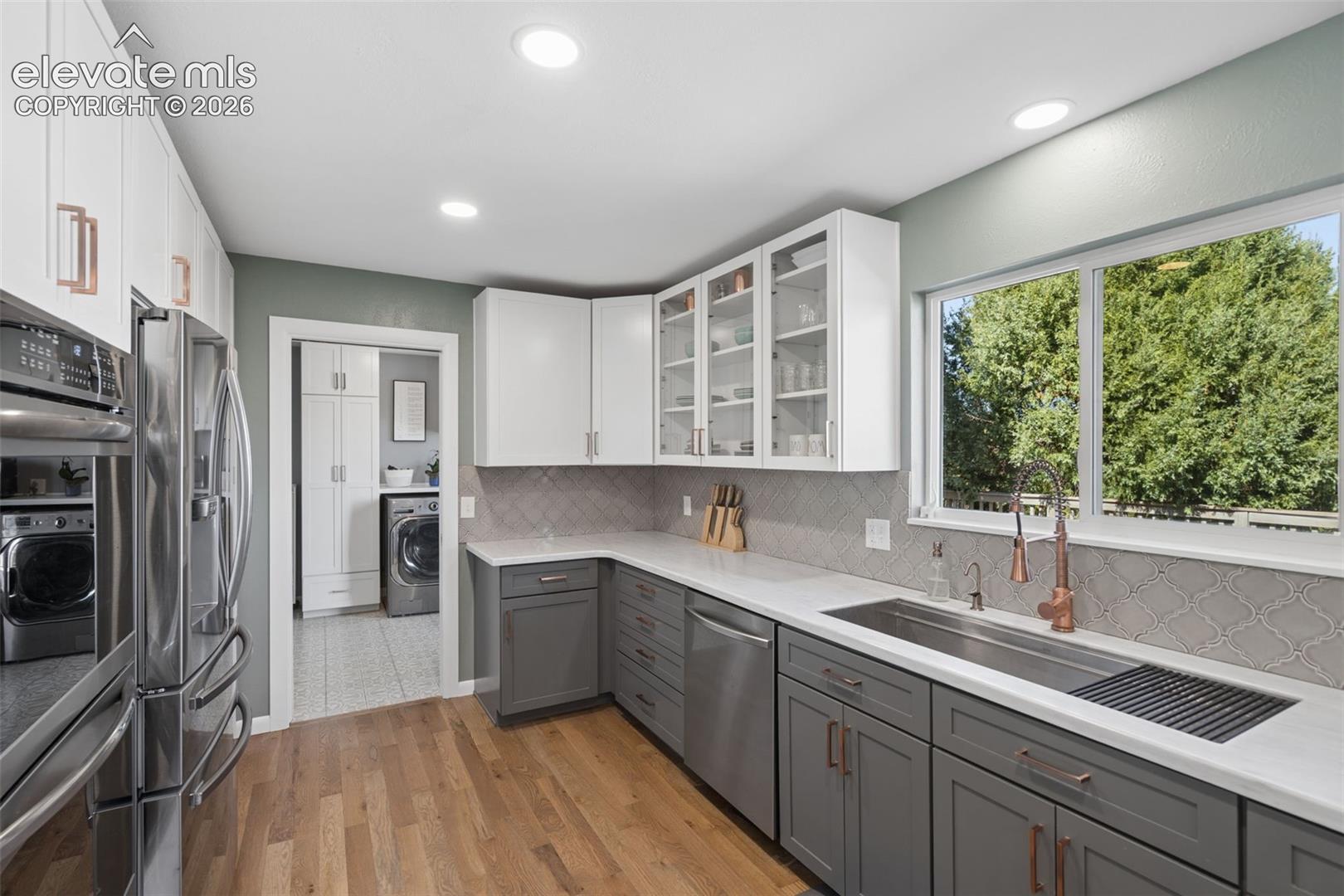 Kitchen featuring white and grey shaker-style cabinetry, white solid-surface countertops, wood-finish flooring, a multi-tonal patterned tile backsplash, and a metallic finish gooseneck faucet