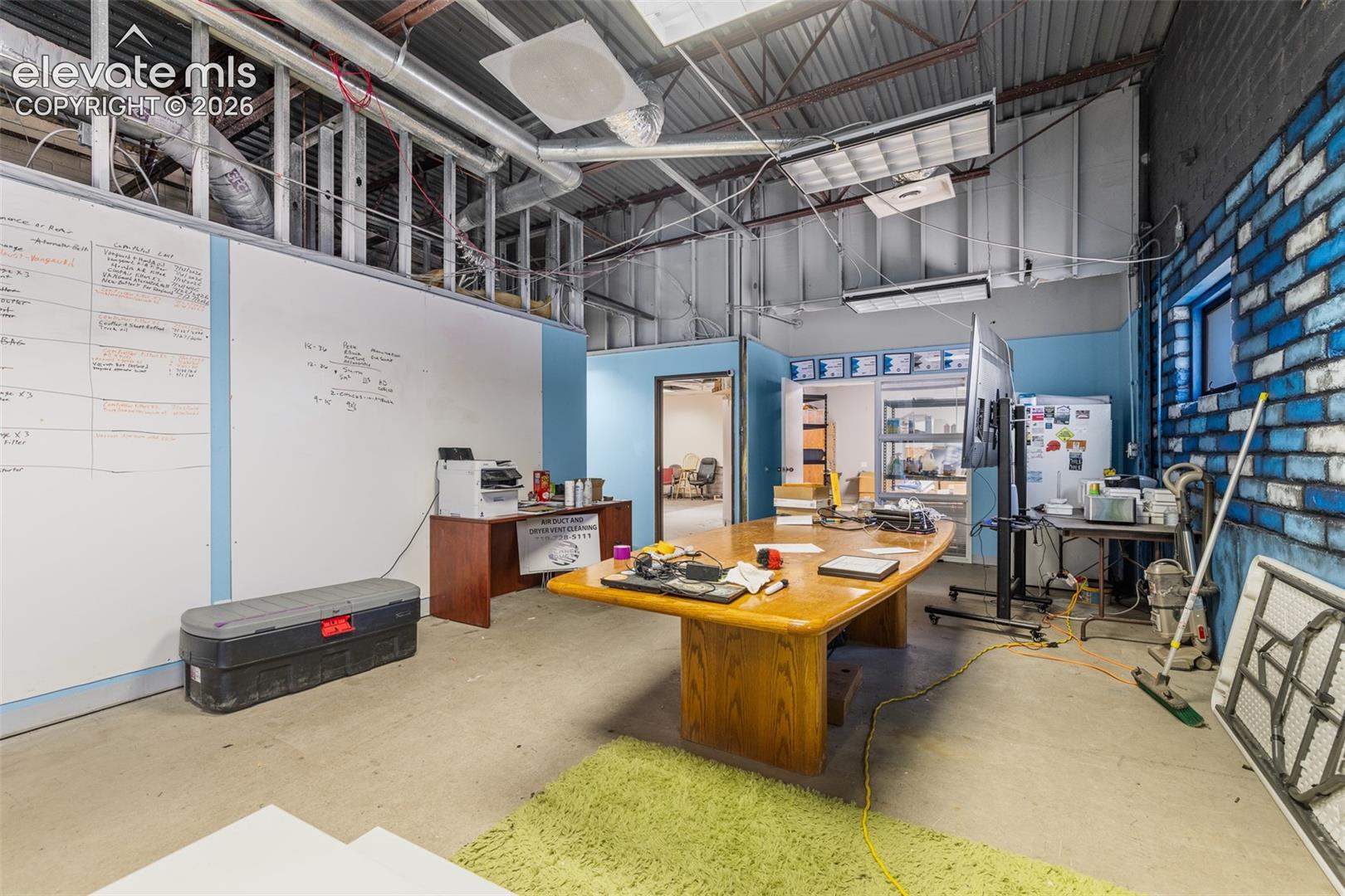 Industrial-style workspace featuring exposed ceiling joists, high-contrast brick accent wall, concrete flooring, and an integrated wall-mounted whiteboard surface
