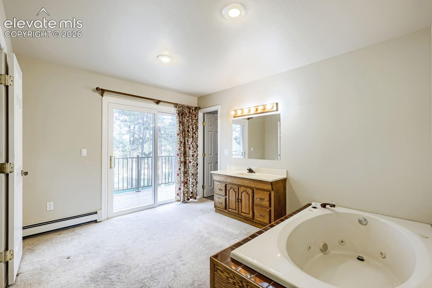 White soaking tub with built-in jets, wood vanity with a single basin, wall-mounted mirror with overhead lighting, and sliding glass door with balcony access