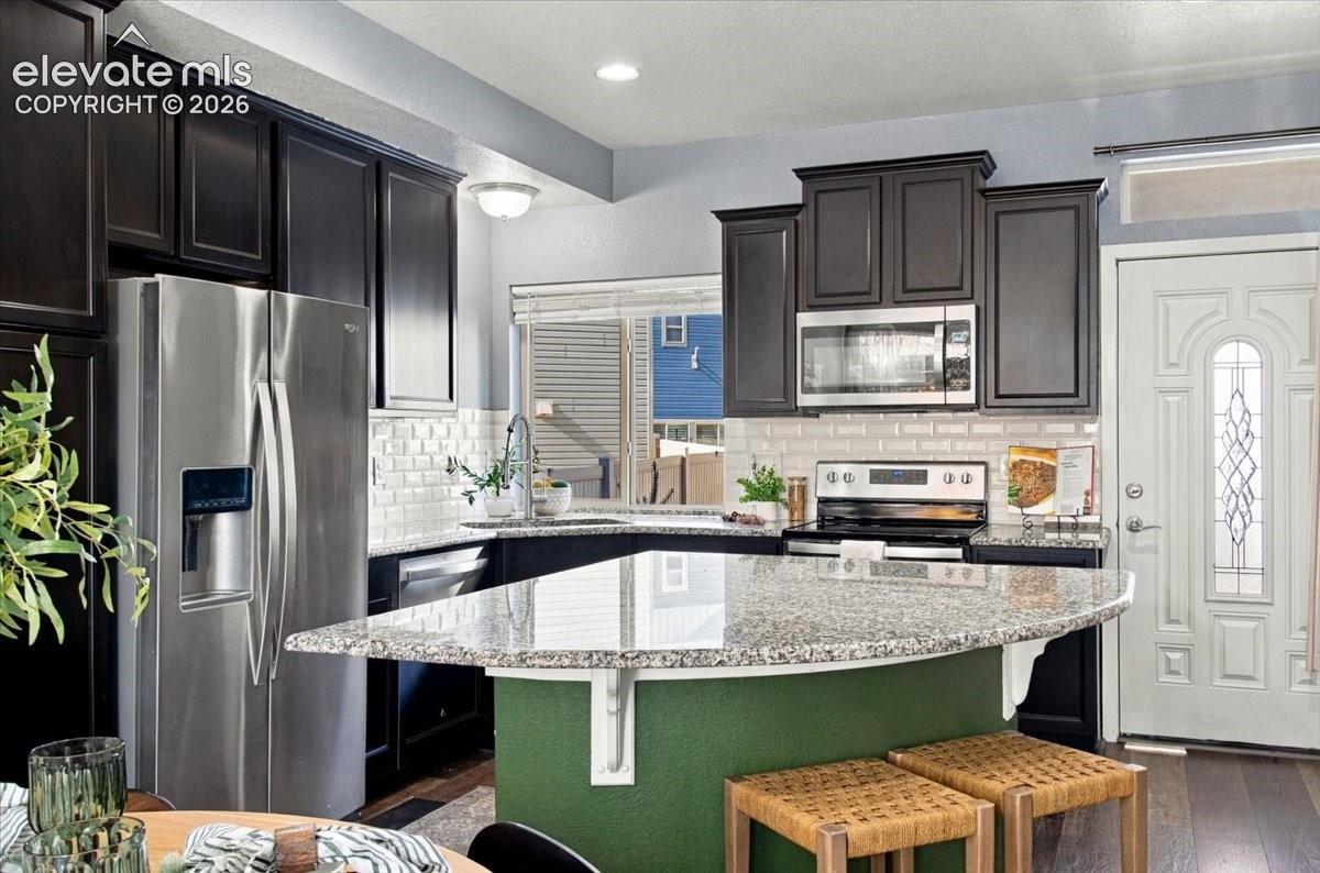 Kitchen featuring dark wood cabinetry, stainless steel appliances, a curved island with a granite countertop, and subway tile backsplash